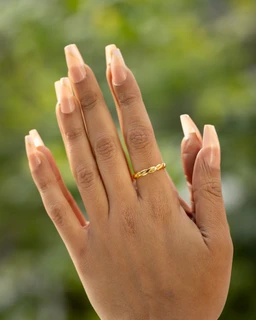 - Description: A single delicate gold-toned ring worn on the middle finger of a hand with neatly manicured nails. The ring features a simple twisted or braided band design.
- Audience and styling: Appears designed for women; minimal, elegant everyday wear suitable for formal or casual looks.
- Materials and color: Gold-tone metal with a warm yellow-gold color; lightweight, slender band.
- Visible attributes: One-layer ring, twisted/braided texture, worn on a clean, natural hand against a blurred outdoor background.