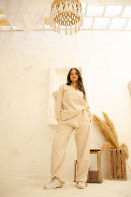 - What the product is: A matching loungewear set (top and bottoms) in a soft beige crinkle fabric.
- Apparent audience: Women, based on the model and styling.
- Notable attributes visible: Light beige/cream color, relaxed fit, long-sleeve button-up top with a tie at the waist, loose-fitting pants, and casual sneakers. The fabric appears crinkled with a soft, cozy texture; the overall look is minimalist and comfortable.