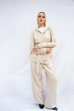 A beige belted loungewear set worn by a female model. The outfit includes a structured zip/knit top with a belt at the waist and matching wide-leg pants; the look is finished with a light outer layer over a white top. Apparent materials suggest soft knit or fleece with a ribbed texture; colours are neutral beige, giving a minimalist, cozy, and casual chic style. Intended audience appears to women's fashion.