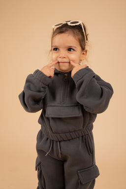 - A young child model wearing a matching gray tracksuit set consisting of a pullover hoodie with a front pocket and matching jogger pants with an elastic waistband and pockets. The outfit appears unisex, suitable for boys or girls, based on style and sizing cues from the model.
- Notable attributes: matte grey fleece material, oversized yet cozy fit, gathered cuffs and waistband, high-neck zip detail on the hoodie, and cargo-style pockets on the pants. The look is casual, sporty, and comfortable for everyday wear.