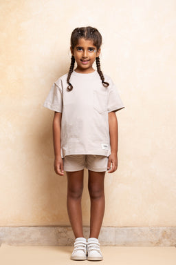 - Product: A unisex oversized short-sleeve T-shirt in beige paired with light beige shorts; the model is a young child.
- Audience and styling cues: Appears designed for kids with a relaxed, gender-neutral fit; light, neutral tones give a minimalist, casual look.
- Notable attributes visible: Soft beige fabric, short sleeves, chest pocket detail on the T-shirt, matching beige shorts, white sneakers. No brand logos visible.
