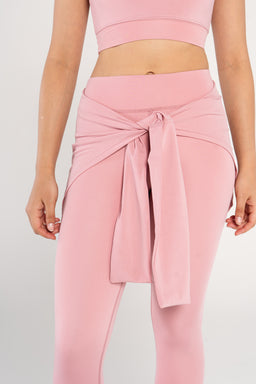 - What it is: A pastel pink waist wrap worn tied at the front over high-waisted leggings, shown on a cropped sports top.
- Audience: Women's (appears styled for adults with a feminine fit and tied sash at the waist).
- Materials/colors/styles visible: Soft pink knit/jersey fabric with a long fabric sash tied at the waist, high-waisted leggings style, cropped top in matching pink. Clean, athletic-loungewear aesthetic.