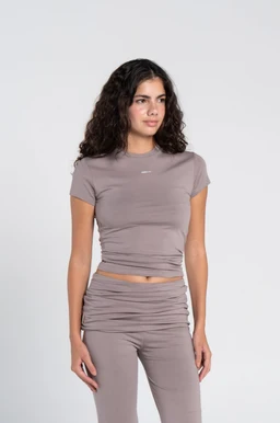 - Product: A cotton short-sleeve crew-neck top with a matching ruched cap-sleeve set (two-piece look: fitted tee and high-waisted ruched leggings).
- Audience: Women (based on slender female model, cut and styling).
- Notable attributes: Soft taupe/greige color, round neck, short sleeves, close-fitting cotton fabric, ruched detail along the midsection and matching high-waisted bottoms, clean minimalist style.