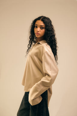 - Description: A loose, oversized beige polo or shirt with dropped shoulders and long sleeves, worn by a woman. The garment has a simple, minimal design with a classic collar and button placket, and the hem appears slightly shorter in front with a curved back.
- Audience and styling cues: The model appears female, suggesting the item is oriented toward women. The relaxed fit and neutral beige color give it a casual, minimalistic streetwear vibe.
- Notable attributes: Light tan/beige fabric, soft drape, long sleeves with wide cuffs, collared neckline, and a clean, contemporary silhouette. The look is understated and versatile for casual outfits.