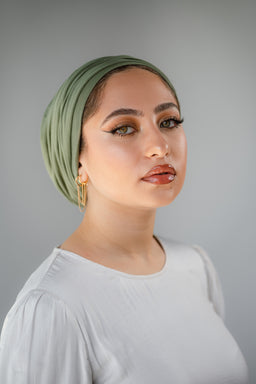 This is a headwrap/headscarf styled as a fashion accessory, shown on a female model. It appears to be a soft, lightweight fabric in a pickle green color, wrapped in a layered turban style that covers the back of the head and a portion of the forehead. The overall look is simple and elegant, pairing with a neutral white top and gold hoop earrings, suggesting a unisex-to-feminine fashion accessory with a smooth, matte finish.