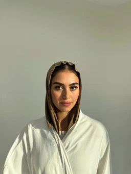 - A light, taupe-toned scarf is draped over a model’s head like a hood, with the fabric wrapped around the sides and tied under the chin in a simple, thin style. The model wears a white top, presenting a clean, minimalist look suitable for unisex or women’s styling.
- Visible materials appear smooth and soft, with a matte finish in clay brown tones; the scarf’s color sits between beige and brown, giving a warm, earthy impression. The overall style is modest and versatile, suitable for casual or layered outfits.