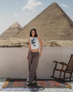 - Product: A white sleeveless tank top with a round neckline, featuring a graphic print on the front.
- Audience: Women (adult female model).
- Visible attributes: White fabric, cropped length showing midriff, graphic black print, paired with loose gray wide-leg pants; casual, boho-style setting with pyramids in the background, suggesting a relaxed, travel-inspired look. No branding visible.