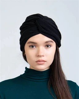 This is a portrait-style product photo showing a woman wearing a black turban-style headwrap with a prominent knot twist. The audience appears to be unisex or women, judged from styling and fit. Notable details: solid black turban, soft fabric with a wrapped, layered design, paired with a dark teal turtleneck. The overall look is modern and minimalist with a matte finish.