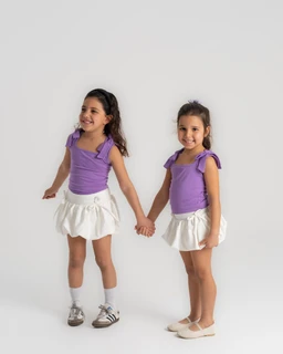 This is a matching pair of girls wearing a purple bow-tie top. The tops are sleeveless with a gathered shoulder strap detail and a small bow on the shoulder, paired with white, ruffled mini skirts; both looks appear coordinated for a unisex or girls’ audience. Notable attributes: bright purple knit fabric, soft pastel white skirts with ruffles, and casual flat footwear (socks with sneakers on one model and ballet flats on the other).