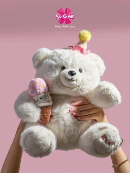 - Product: A fluffy white teddy bear holding a small ice cream cone, with a pink party hat on its head. The bear is being held by a person, showing its soft, plush texture and round facial features.
- Intended audience: Unisex, with a playful, cuddly design likely aimed at children and collectors.
- Notable attributes: Soft, plush fur in white; light pink accents on the hat and the ice cream cone; black stitched eyes and nose; pink background; small heart detail on the bear’s foot. The branding “SUGAR” is visible near the top, but no specific brand name is identified.