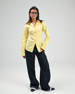 This is a pale yellow button-down chemise with a relaxed, tailored fit. The model (adult woman) wears it with dark wide-leg jeans and heeled shoes, giving a chic, casual-office vibe. Notable attributes include the lightweight shirt fabric, long sleeves, collar, and front button closure with a slightly curved hem.