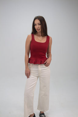 A sleeveless, fitted tank top in dark red with a peplum ruffle at the hem. The model appears to be a female, styled with white high-waisted wide-leg pants. Notable details include a square neckline, smooth knit fabric, and a slight stretch finish, presenting a casual yet dressy look in a solid rich red color.