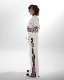 - What it is: A metallic tracksuit set consisting of a long-sleeve top and matching wide-leg pants with a shiny metallic panel accent.
- Audience: Appears designed for women (fit and styling on a female model).
- Notable attributes: White base fabric with a lavender metallic stripe/panel along the arms and down the outer leg; relaxed fit top with a cropped or boxy silhouette and cuffed or elasticized waist, full-length pants with a vertical metallic stripe; glossy metallic finish; minimalist, sporty-urban style.