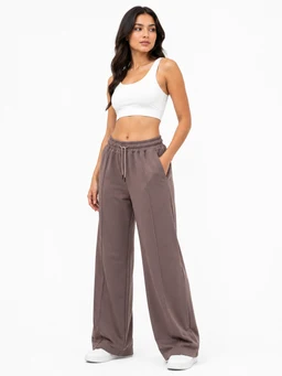 These are women's wide-leg trousers styled for a relaxed, casual look. The model wears a white crop top to pair with the high-waisted, loose-fitting pants that have a drawstring waistband and side pockets, creating a comfortable, athleisure vibe. Fabric appears lightweight with a soft drape in a muted taupe/stone color; overall silhouette is flowy and floor-skimming, with a Hashira fabric texture suggested by the smooth, matte finish.
