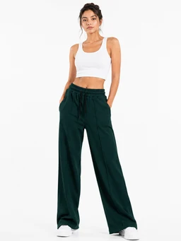 - Description: A female model wearing wide-leg trousers with a relaxed, high-rise fit and a drawstring waist. She pairs them with a white crop top, showing a casual, athleisure vibe.
- Audience and styling: Female (women’s) audience; casual, sporty-luxe styling with a minimal, clean look.
- Visible attributes: Dark green (deep emerald) wide-leg trousers in Hashira fabric; drawstring waistband; front pockets; lightweight, fluid drape; white sneakers and a white crop tank top as styling. No branding visible.
