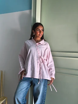 - Product: Side Bow Linen Shirt in Soft Pink
- Audience: Women's (model styling and fit suggest feminine silhouette)
- Notable attributes visible: lightweight, loose-fit linen shirt in soft pink, with a side tie/bow detail at the waist, long sleeves, a collared neckline, and a relaxed, slightly oversized cut. Paired with blue jeans. The shirt appears breathable and textured typical of linen.