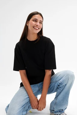 Product is an oversized black short-sleeve T-shirt. Appears modeled by a woman (adult female) for unisex/women’s sizing with a relaxed, oversized silhouette. Visible attributes: solid black knit fabric, crew neck, loose fit through the body and sleeves, paired with light blue wide-leg jeans for styling; no branding visible.