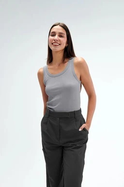 A sleeveless grey tank top with a scoop/rounded neckline. The model appears to be an adult woman, suggesting a women's cut and fit. Visible attributes: solid light grey fabric, close-fitting sleeveless style, paired with high-waisted dark grey/black trousers. The overall look is casual, with a simple, minimal design and a soft matte finish. Key materials and styling cues: knit or jersey fabric, plain colour, basic tank top silhouette.