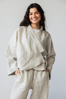 - What the product is: A light, draped cardigan-style top with an attached scarf-like tie, paired with matching relaxed trousers. The set appears to be a coordinating loungewear or casual outerwear look.

- Apparent audience: Women (model is female; styling is relaxed and oversized, typical of women's loungewear).

- Notable attributes: Raw beige/neutral fabric in a soft, textured linen-like material. Loose, oversized fit with wide sleeves and layered, wrap-front scarf detail. Matching wide-leg trousers with an elastic or drawcord waist. Unstructured, minimalist style with a calm, neutral color palette.
