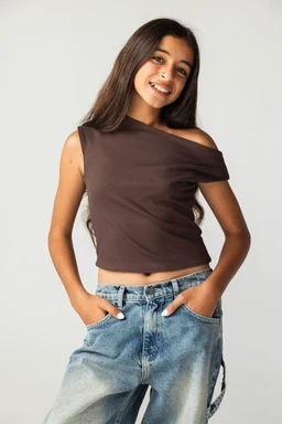 Product: Asymmetric Brown Tank Top - Kids
- Audience: Kids (girl) based on styling with slim fit and casual look, mid-length hair model, and cropped top.
- Visible attributes: One-shoulder asymmetrical neckline with a brown solid color, sleeveless design, cropped length, soft/stretchy knit fabric, paired with light-wash denim jeans. Minimal seams and clean edge hem, no brand logos visible.