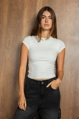 - What the product is: A short-sleeve white ribbed tank top for youth, shown as a fitted cropped tee. 
- Intended audience: Youth girls, based on the model and sizing/fit.
- Notable attributes visible: White color, slim fitted cut, short sleeves, crew/round neckline, cropped length, and lightweight stretch fabric. The top is paired with dark jeans for a casual, modern look.