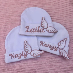 - Product: A set of white baby hats with matching folded cuffs featuring embroidered names and wing accents; the visible items include a beanie-style hat and two sleeve/ear-cover cuffs with name embroidery and small wing motifs. 
- Intended audience: Infants (unisex appearance based on styling and size).
- Notable attributes: Soft white fabric, light brown embroidery, cursive name lettering (e.g., Lalid, Nazly, Kenzy) with embroidered wings on each piece; simple, cozy knit style suitable for babies.