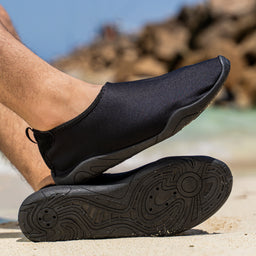 - What the product is: A black slip-on water-sock / minimalist shoe with a neoprene-like upper and flexible sole, shown being worn with one foot resting on the sole.
- Apparent intended audience: Unisex (suitable for both men and women), judging from the neutral black color, streamlined slip-on design, and casual outdoor style.
- Notable attributes visible: Matte black fabric upper, low-cut ankle, pull-tab at the heel, thick black rubber sole with a wavy tread pattern, and a sleek, sock-like silhouette suitable for light walking or beachwear.