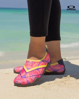 - A pair of pink swim/athletic shoes with a lightweight, flexible sole, shown on a beach. 
- Appears designed for women or girls based on the styling and cut, with a bright pink and yellow pattern and a low-cut, slip-on design. 
- Notable features include a colorful fabric upper with abstract/spiral prints, yellow piping accent, grey/neutral sole, and a breathable, beach-ready look.