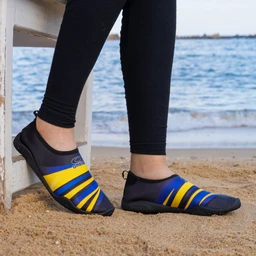 This photo shows a pair of water shoe/barefoot shoes worn by a person standing on sand at the beach. The apparent audience is unisex, judged from the simple, aquatic-sport styling and neutral fit. Notable attributes: dark navy upper with bright yellow and blue diagonal stripes, cut-low profile with a slip-on design and a small loop at the heel; flexible, grippy sole suitable for wet sand or water activities. The model is wearing black leggings, and the scene is outdoors by the water.