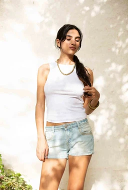 - What the product is: A sleeveless tank top in white, shown on a female model with light-wash denim shorts. The top is fitted with a crew/round neckline and exposes the midriff.
- Apparent intended audience and styling: Women or girls (adult female model); casual summer or warm-weather look. Model accessorized with a chunky chain necklace and a bracelet, with hair in a side ponytail.
- Notable material, color, and style attributes: Solid white knit jersey fabric; cropped length; ribbed/soft stretch finish; simple sleeveless cut with wide shoulder straps and high armholes; paired with light blue distressed denim shorts.