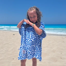 A kids’ poncho in a vivid blue maze-like geometric print, shown on a young girl at the beach. The gusty, oversized poncho style with short sleeves suggests a unisex or girls’ summer cover-up, intended for playful, water-friendly use. Visible features include a bold blue-and-white pattern, loose drape, and a soft towel-like fabric with a small tag at the lower front.