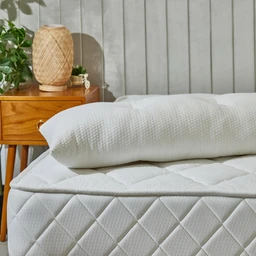 - What the product is: A white bed pillow set arranged on a mattress, with a matching rolled pillow and a quilted cover.
- Apparent audience: Unisex (suitable for men and women) based on the neutral, classic bedding style.
- Notable attributes visible: White quilted fabric with a diamond pattern on the mattress cover, a soft-textured woven pillow, and a small wooden nightstand with a decorative wicker lamp in the background; overall style is clean, cozy, and restful. Material hints include cotton or cotton-blend quilted fabric and a textured pillow cover.