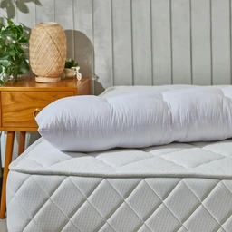 - What the product is: A long, cylindrical pillow laid across a bed, paired with a white quilted mattress cover.
- Apparent audience: Unisex (suitable for both men and women), judged by neutral styling and standard long pillow shape.
- Notable attributes: White fabric with a smooth, quilted surface; long tubular shape likely used for body support; appears to be a plush or microfiber fill, giving a softly rounded, fluffy look. The setting shows a wooden nightstand and a woven lamp for a cozy bedroom style.
