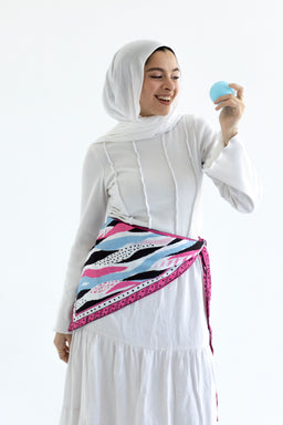 This product shows a woman wearing a white ensemble with a hijab, holding a small blue ball, suggesting a playful fashion look. The outfit is a long-sleeve, ankle-length white dress and a light-weight scarf tied around the waist as a belt or sash, in a bold, multicolor patterned scarf with pink borders. The visible materials appear to be soft, breathable fabrics (cotton or jersey) for the dress, with a lightweight, printed scarf featuring pink, black, white, and blue wave-like patterns. The overall style is modest, sporty-chic, suitable for casual or active daily wear. Intended audience: women's. Notable attributes: white color base, scarf belt with vibrant waves pattern, pink trim, hijab styling, relaxed fit.