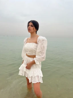 A white, two-piece set featuring a cropped, smocked bandeau top and a matching tiered mini skirt. The long puff sleeves add drama while remaining lightweight and airy, perfect for a beach or resort look; the fabric appears to be eyelet or broderie Anglaise with small embroidered holes. The overall style is feminine and romantic, intended for a women's audience.