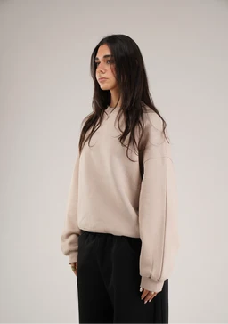 - A beige crewneck sweater modeled by a woman, with a relaxed, oversized fit and drop shoulders. The fabric appears soft and slightly textured, in a light taupe-beige color, paired with black wide-leg pants for contrast. The overall look is casual, aiming at a women's audience, with visible features: long sleeves with ribbed cuffs and a ribbed hem.