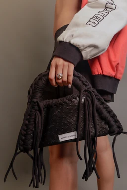 - What it is: A small black handbag with a braided fringe detail and drawstring sides, held by a model.
- Intended audience: Unisex with a chic, edgy vibe; appears styled for women given the handbag carry and ring detail.
- Notable attributes: Black fabric with braided fringe accents, woven texture, drawstring closure, compact rectangular shape, and a small label visible on the front. The model is wearing a color-block jacket with sporty lettering, suggesting a casual to streetwear style.