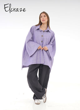 A light purple linen shirt with an oversized, relaxed silhouette. The model appears to be a woman, wearing the shirt as a loose button-front top with wide sleeves and a hem that falls around hip length; the shirt has a pointed collar and large front buttons. Visible material is a textured linen in a soft lavender shade; styling is casual and flowy, paired with dark wide-leg pants and sneakers for a comfortable, effortless look.