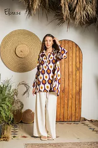 - What the product is: A semi-formal women’s outfit set featuring a loose, patterned tunic or shirt paired with wide-leg pants.
- Apparent audience: Women (unisex not applicable), based on the model, styling, and fit.
- Notable attributes: The top has a bold purple and brown circular pattern on a light background, with a relaxed, knee-length tunic silhouette. The bottoms are wide-leg, cream-colored pants. The overall look is casual yet semi-formal, with natural straw hat accessory and an earthy, boho vibe. Materials and textures: lightweight fabric for the top and flowy pants, colors include purple, brown, and cream.
