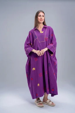 - Product: A loose, ankle-length purple dress with a relaxed fit and oversized silhouette. It features a collar, a short zippered placket, and wide sleeves, giving a kaftan-like look. The fabric is decorated with small, colorful embroidered motifs scattered across the front and sides.

- Audience: Women (unisex styling but presented on a female model).

- Notable attributes: Bright purple color, soft, flowy fabric, mid-length to long dress, casual yet bohemian style with embroidery and relaxed drape. Worn with flat sandals for a laid-back, comfortable vibe.