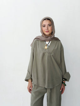 - A lightweight, loose-fitting tunic and matching pants set in a muted mint/sage green. The top features a relaxed silhouette with a front seam detail and wide, long sleeves that have gathered cuffs; the bottoms are full-length, straight-cut pants. The model wears a neutral-toned hijab and a geometric pendant necklace, suggesting a modest, contemporary lounge or casual wear style. Notable materials appear soft and flowy, with a matte finish suitable for comfortable everyday wear.