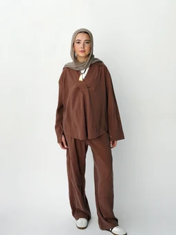 The photo shows a brown, loose-fitting two-piece set consisting of a long-sleeve top and wide-leg pants. The model wears a matching hijab, giving a modest, unisex to women’s casual/relaxed look. The fabric appears soft with a matte finish in a warm brown tone; the top has a relaxed silhouette with a slight drape and a split neckline, while the pants are high-waisted with a relaxed, straight leg. No branding visible.