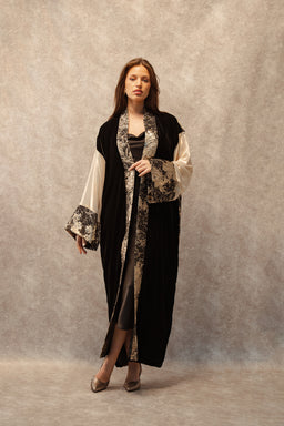 - What the product is: A long, draped cardigan or robe-style outer garment with wide sleeves and a contrasting patterned trim, shown worn open over a black top and light-colored pants.
- Apparent audience: Women (based on the model and styling).
- Notable attributes: Velvet-like black main fabric with light, floral-patterned cream trim along the edges and cuffs; floor- to ankle-length cut; wide kimono-style sleeves and an open front; pairing suggests a formal or dressy, bohemian-inspired look.