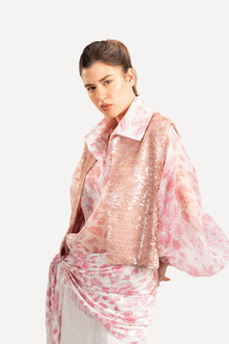 - A pink organza top with a shiny, sheer finish is styled over a light pink patterned dress or top. The model wears a cropped, soft pink sequin-like bolero or shrug layered over the outfit, creating a delicate, feminine look.
- Audience: women’s
- Notable attributes visible: sheer pink organza fabric, light pink/rose color palette, sequined or metallic-textured outer layer (bolero or short jacket) in a pale pink, collar and loose-fitting top styling with flowing, lightweight materials.