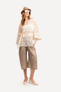 - A two-piece matching set featuring a light, airy lace top and a pair of knee- to mid-calf length wide-leg Bermuda shorts in metallic beige/taupe tones.
- Appears styled for women; model wears a wide-brim hat, large sunglasses, and pointed flats, suggesting a casual, boho-chic daytime look.
- Visible materials and colors: sheer lace overlay and lined bodice in pale champagne/metallic beige, with loose, flowy sleeves and a sash tie at the waist; shorts have a soft, drapey fabric with a wide, cropped silhouette.
