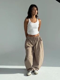 - What the product is: A pair of plaid pants in brown tones, styled with a relaxed, loose fit and gathered waistband.
- Apparent audience: Unisex or women’s, based on the model and fit (female-presenting with a crop top and relaxed silhouette).
- Notable attributes visible: Brown plaid pattern, wide, harem-style or balloon-leg silhouette, elasticized waist with a drawstring or elastic, cropped length near the ankle, light fabric with a soft drape, paired with a white ribbed tank top and casual sneakers.