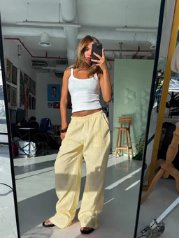 - What the product is: A pair of yellow striped linen pants, shown on a female model in a bright studio setting.
- Intended audience: Women's (fashion-forward relaxed-fit pants on a female model).
- Notable attributes visible: Light yellow linen fabric, wide-leg relaxed silhouette, elastic or drawstring waistband, high-rise fit, casual summer style. The model pairs the pants with a white sleeveless crop top and flat sandals, emphasizing a breezy, warm-weather look.