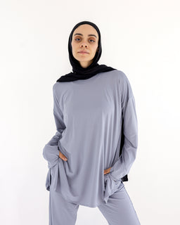 - This photo shows a modest, loose-fitting two-piece lounge set in a soft lavender-grey color, consisting of a long-sleeve tunic top and matching wide-leg pants. The model is wearing a black hijab, and the outfit has a relaxed, comfortable silhouette with pockets visible on the tunic sides. Notable materials and styling cues include a drapey, jersey-like fabric and a monochrome look with a simple, understated finish. Audience: women.