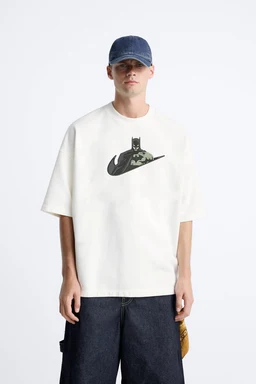 A short-sleeve unisex or men's T-shirt in a light/cream or white color featuring a large Batman-themed embroidery on the chest area, with a dark graphic over a circular or swoosh-like background (similar to a stylized emblem). The visible styling includes a relaxed fit, dropped shoulder, and paired with a denim cap and dark jeans for a casual streetwear look; material appears to be a medium-weight cotton jersey. Notable attributes: mid-weight cotton fabric, light color, oversized/boxy silhouette, and a single embroidered graphic at front.