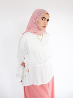 - What it is: A satin evening blouse in white, styled with a soft, flowing silhouette.
- Intended audience: Women (modest, with headscarf shown; hijab-friendly styling).
- Notable attributes: White satin-like fabric with a loose top and elasticized waist peplum effect, long sleeves, and a light pink hijab. Paired with pink bottoms in the photo. Features a simple layered jewelry look. No visible branding.